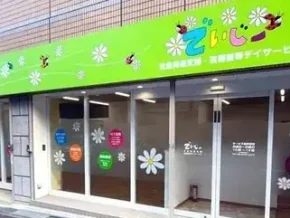Front of a brightly colored storefront with a lime-green sign and white daisy window decals; appears to be a local welfare or day service center in Japan.