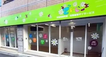 Front of a brightly colored storefront with a lime-green sign and white daisy window decals; appears to be a local welfare or day service center in Japan.