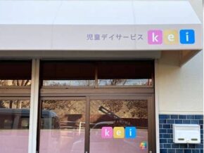 Entrance to a children's day service center with a glass door and a pastel sign in Japanese '児童デイサービス' and colorful 'kei' logo on the window.