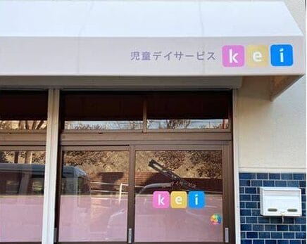 OIP 4 | 保育士求人|大阪・兵庫・関西の保育士・幼稚園教諭の就職転職支援|保育のせかい Entrance to a children's day service center with a glass door and a pastel sign in Japanese '児童デイサービス' and colorful 'kei' logo on the window.
