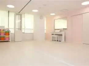 Bright, empty dance studio or classroom with pastel pink walls, mirrors, a white piano against the back wall, and colorful cubbies on the left.