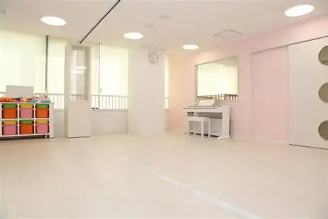 Bright, empty dance studio or classroom with pastel pink walls, mirrors, a white piano against the back wall, and colorful cubbies on the left.