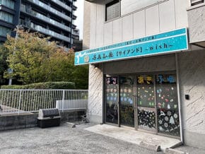 Storefront with a blue sign reading 'Wish' above glass doors covered in colorful decorative stickers; a small tree and metal railing on the left, in a city setting with a high-rise building in the background.