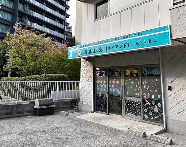 Storefront with a blue sign reading 'Wish' above glass doors covered in colorful decorative stickers; a small tree and metal railing on the left, in a city setting with a high-rise building in the background.