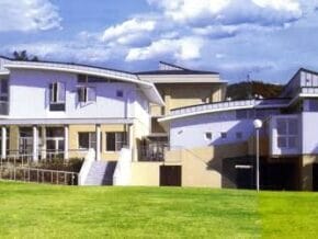 Modern white, multi-wing building with large windows, set on a well-kept green lawn under a blue sky.