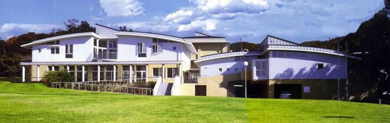 Modern white, multi-wing building with large windows, set on a well-kept green lawn under a blue sky.