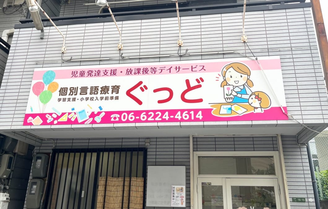 Exterior storefront sign for a children's developmental support and after-school service, with a large red 'ぐっど' and phone number 06-6224-4614, plus cartoon illustrations.