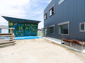 Exterior view of a blue metal building with several windows, a colorful mural with Japanese characters on a covered wall, and a dirt yard with a wooden fence and bench.