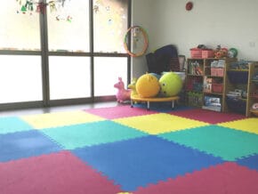 Colorful children's playroom with interlocking foam mats, shelves of toys, a hula hoop, and large windows letting in light.