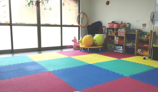 Colorful children's playroom with interlocking foam mats, shelves of toys, a hula hoop, and large windows letting in light.