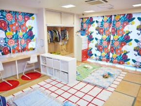 Bright kids' playroom with colorful floral curtains, an open wardrobe, white cubbies, and soft floor mats on a checkered rug.