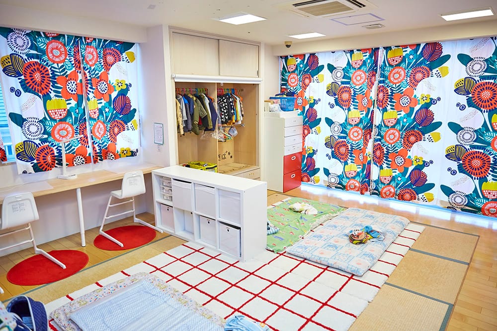 Bright kids' playroom with colorful floral curtains, an open wardrobe, white cubbies, and soft floor mats on a checkered rug.