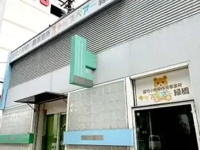 Outside view of a gray building with a teal cross sign and a doorway featuring a bear logo, indicating a children's clinic in Japanese.