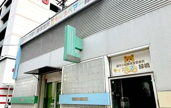 Outside view of a gray building with a teal cross sign and a doorway featuring a bear logo, indicating a children's clinic in Japanese.