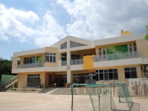 Front view of a two-story yellow and beige school building with a central clock above the entrance and glass doors, adjacent to a dirt playground and green fencing under a blue sky.