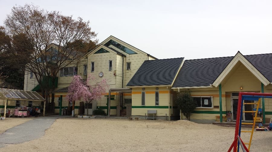 Two-story beige school building with green trim and a dark roof, next to a sandy playground and a pink blossoms tree in front.