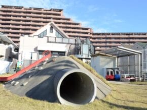 Concrete mound playground with a large tunnel slide and red side rail, set against apartment buildings in the background.