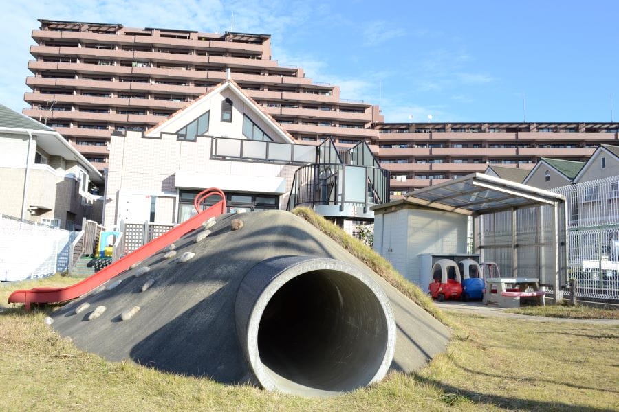 Concrete mound playground with a large tunnel slide and red side rail, set against apartment buildings in the background.