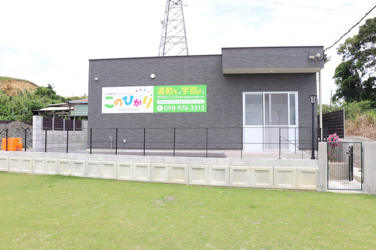 Grey brick school building with a bright, multi-colored sign advertising Akano School, fenced yard, and grassy foreground.