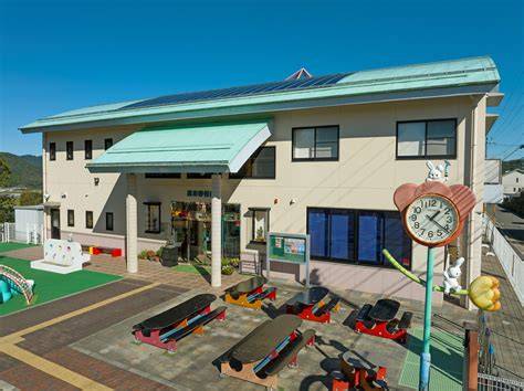 Two-story beige school building with green awnings; courtyard has red and black benches, a small playground, and a large round clock on a pole.