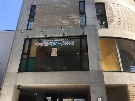 Modern concrete building with a large storefront window displaying a bear mascot and Japanese text, entryway beneath.