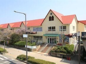 Beige, two-story building with red gabled roofs and a green awning over the main entrance, surrounded by trees and a sidewalked street in front.