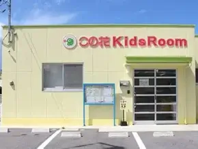 Exterior of a pale yellow building with a red 'Kids Room' sign, glass entrance door, blue framed bulletin board, and a small parking area in front.