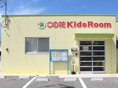 Exterior of a pale yellow building with a red 'Kids Room' sign, glass entrance door, blue framed bulletin board, and a small parking area in front.
