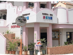 Pink and white kindergarten building with rounded arches, signs in Japanese, and potted plants at the entrance; a cartoon mascot near the doorway.