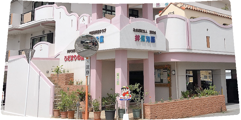 Pink and white kindergarten building with rounded arches, signs in Japanese, and potted plants at the entrance; a cartoon mascot near the doorway.