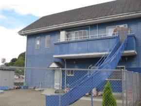 Blue two-story building with an external blue staircase and metal railing, fenced yard, and scattered supplies outside.