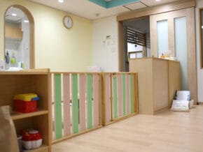 floor01_image02_l | 保育士求人|大阪・兵庫・関西の保育士・幼稚園教諭の就職転職支援|保育のせかい Pediatric clinic reception with a wooden safety rail, pastel green and pink slats, and a wall clock.