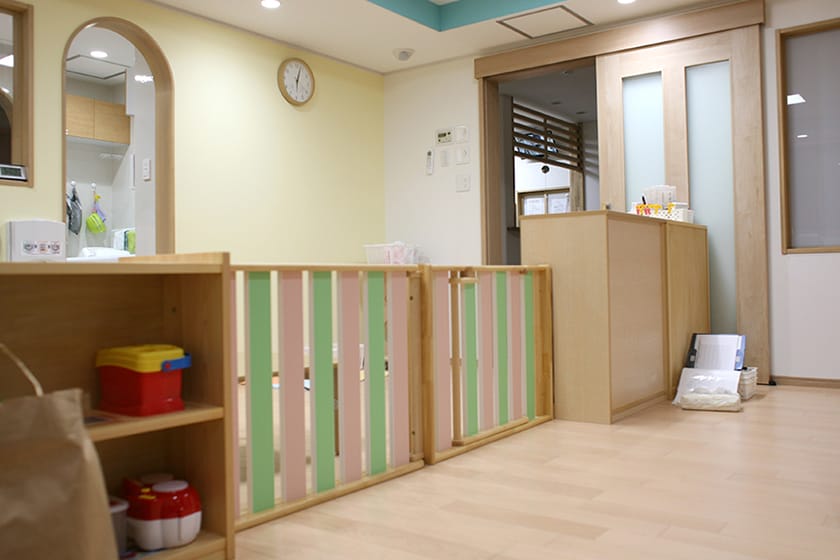 floor01_image02_l | 保育士求人|大阪・兵庫・関西の保育士・幼稚園教諭の就職転職支援|保育のせかい Pediatric clinic reception with a wooden safety rail, pastel green and pink slats, and a wall clock.