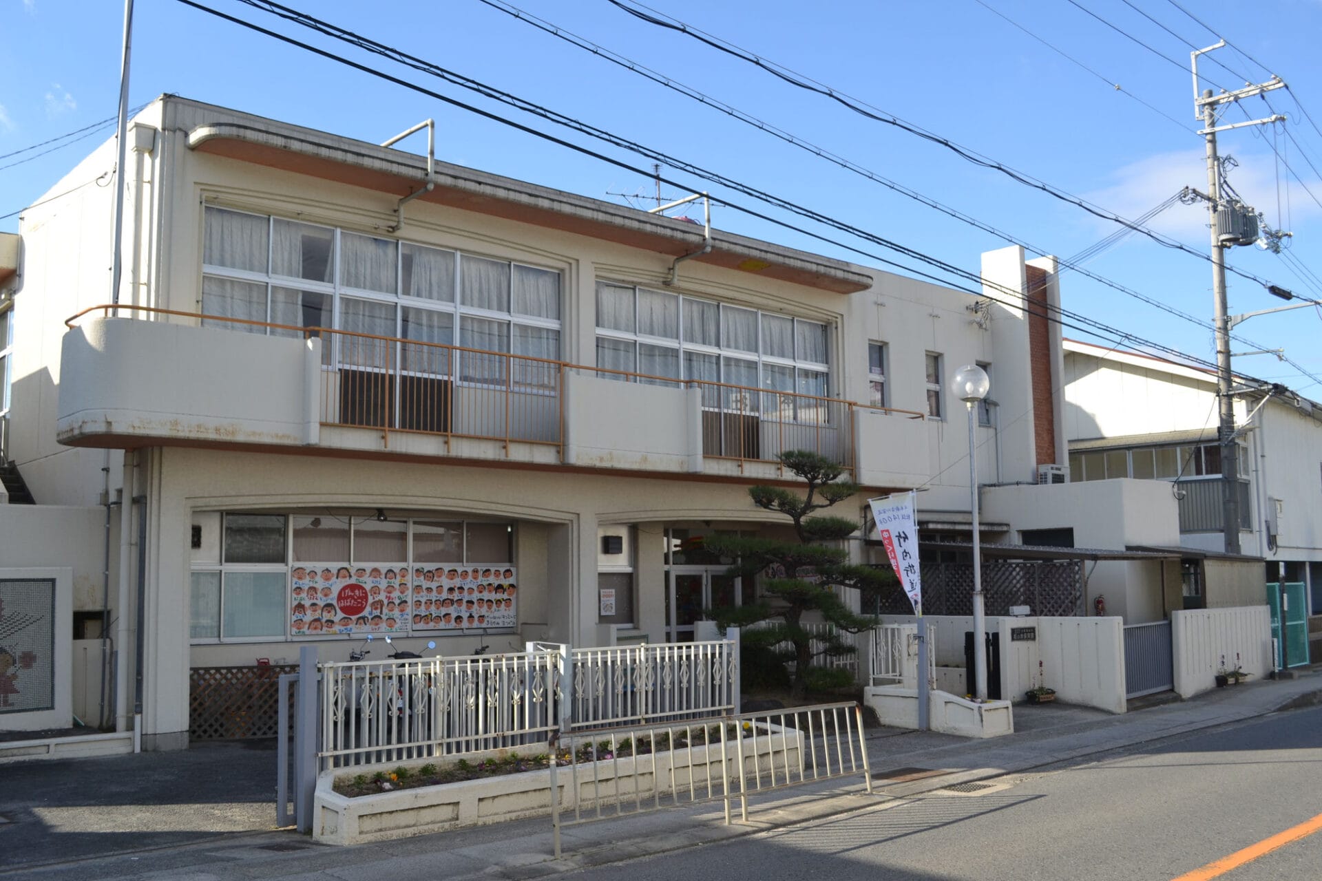 White two-story building with large windows and an orange railing along a balcony, on a street with overhead power lines and a clear blue sky.