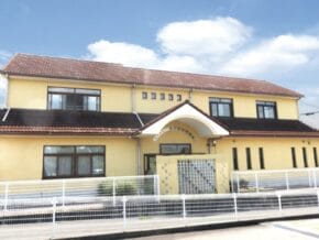 Two-story yellow building with a brown tiled roof, curved white arch over the entrance, and a white metal fence along the street.