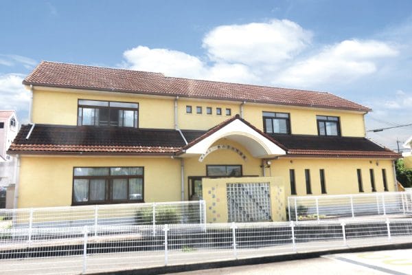 Two-story yellow building with a brown tiled roof, curved white arch over the entrance, and a white metal fence along the street.