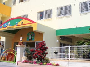 Colorful daycare or preschool exterior with a bright archway, cartoon decorations, and a gated entrance surrounded by plants.