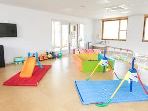 Indoor kids’ playroom with a small orange slide on a red mat and colorful soft play equipment on padded blue mats; bright and welcoming.
