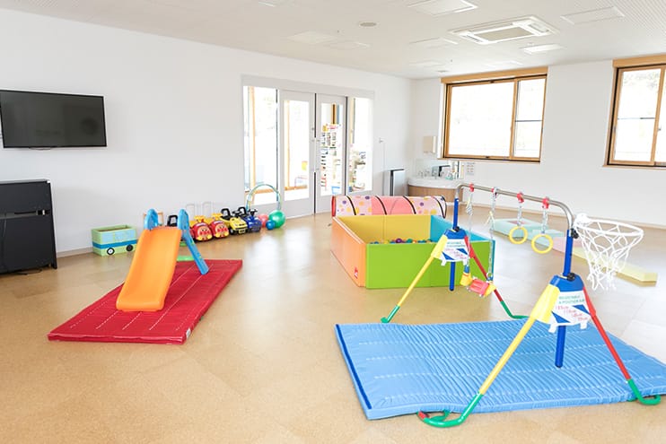 Indoor kids’ playroom with a small orange slide on a red mat and colorful soft play equipment on padded blue mats; bright and welcoming.