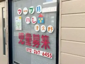 Storefront window advertising child recruitment for a daycare, with red Japanese text, round colorful logos, a poster, and phone number 072-267-6455.
