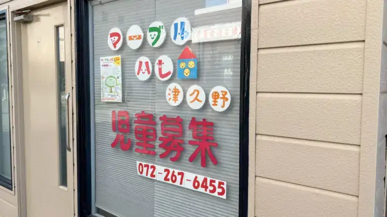 Storefront window advertising child recruitment for a daycare, with red Japanese text, round colorful logos, a poster, and phone number 072-267-6455.