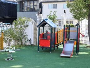 Colorful children's playground with a slide and climbing structure on green turf. Fenced yard and trees in the background.
