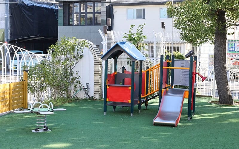Colorful children's playground with a slide and climbing structure on green turf. Fenced yard and trees in the background.