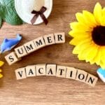 Wooden surface with two yellow sunflowers, scrabble tiles spelling SUMMER VACATION, a straw hat, green leaves, and blue toy dolphins.