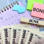 Calendar page open to June with cloud-shaped erasers, a sticky note reading BONUS, a wooden 'JUNE' block, and fanned 10,000 yen banknotes.