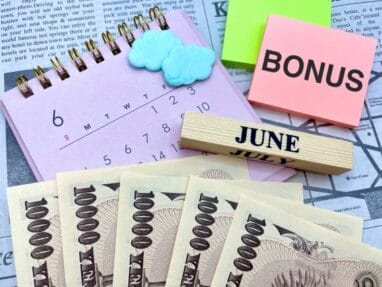 Calendar page open to June with cloud-shaped erasers, a sticky note reading BONUS, a wooden 'JUNE' block, and fanned 10,000 yen banknotes.