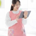Young woman in a pink apron uses a tablet in a bright, sunlit room (kitchen area).