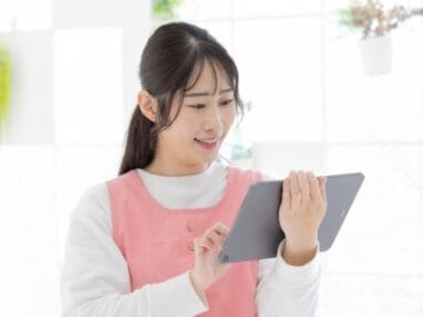 Young woman in a pink apron uses a tablet in a bright, sunlit room (kitchen area).