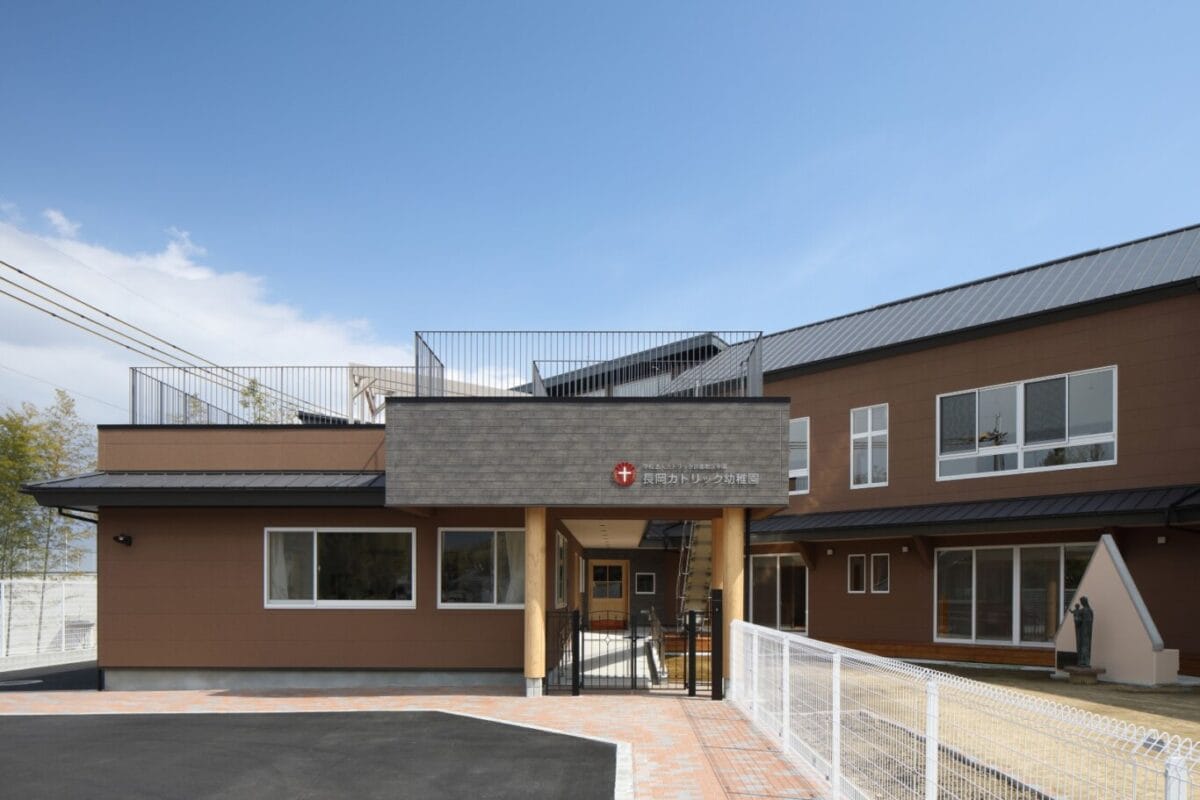 Modern brown two-story building that appears to be a kindergarten or daycare, with a covered entrance supported by wooden columns and a fenced walkway.