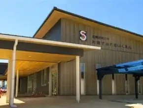 Exterior of a single-story public building with a tan facade, a sloped roof, and a covered entrance; Japanese signage near the top.